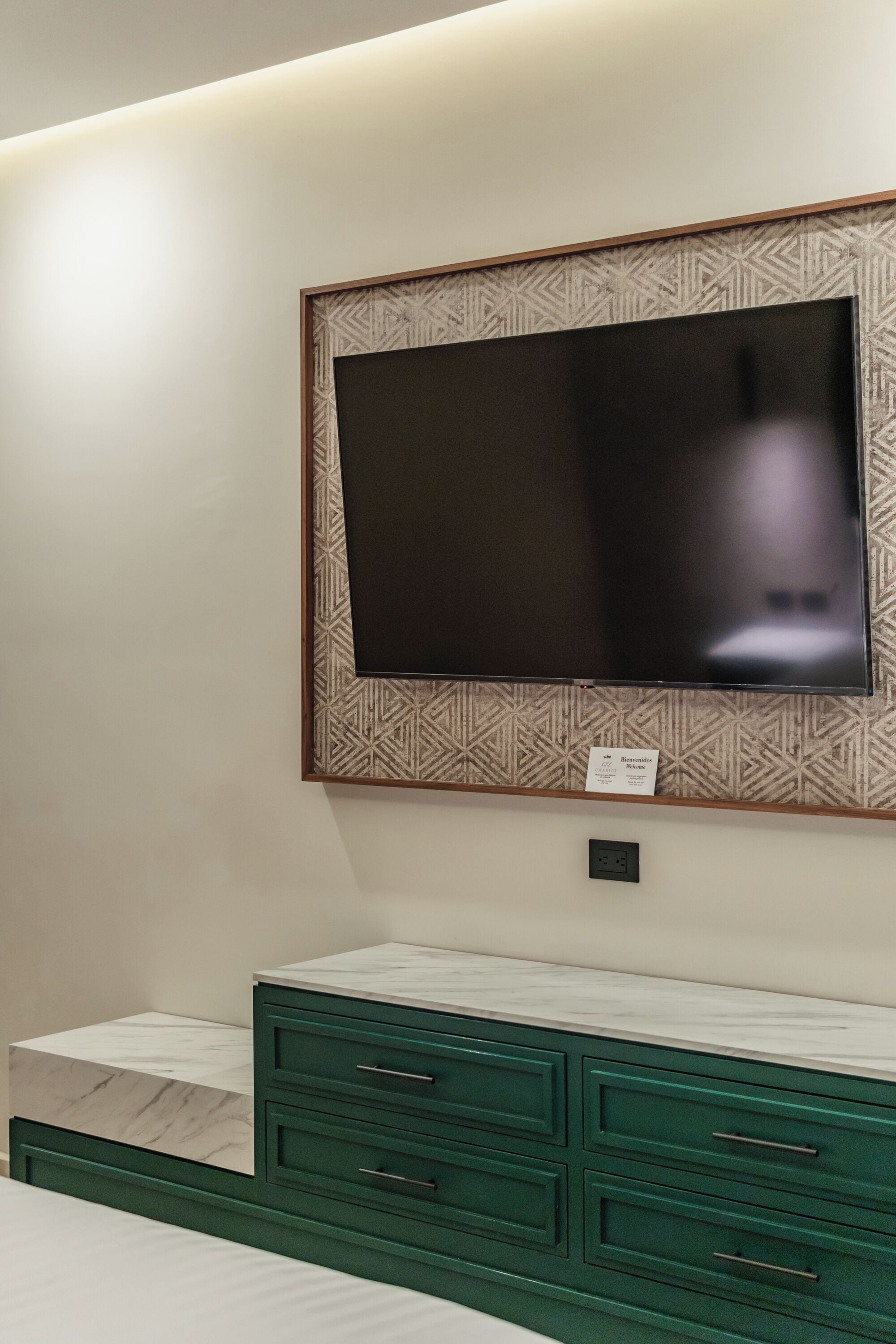 A bedroom with a large flat-screen TV mounted on the wall, a green dresser with drawers, and a marble accent piece on the dresser.