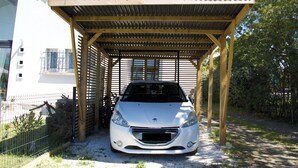 En bil parkeret under en træpergola med et metal tag, ved siden af en hvid bygning med balkon.