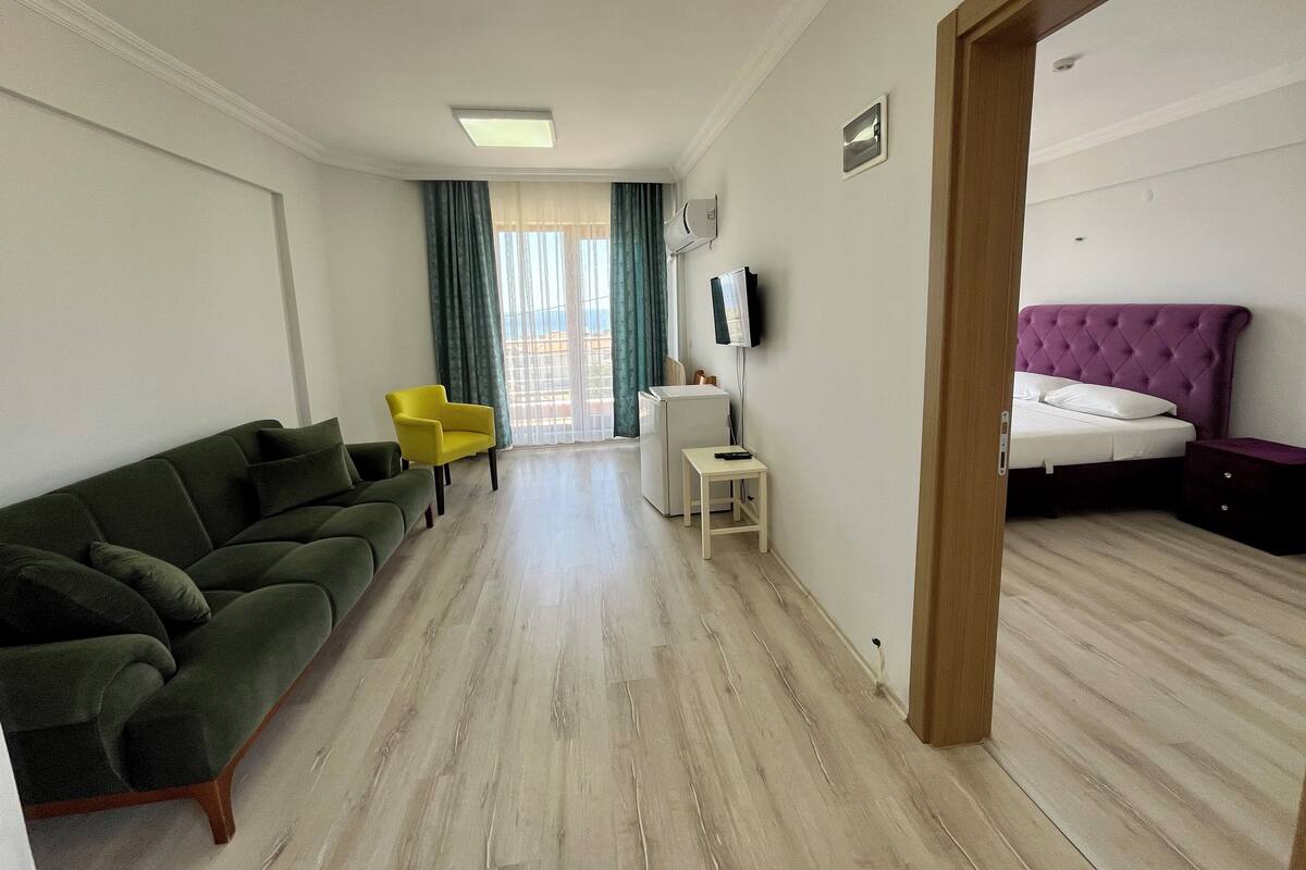 A hotel room with a green sofa, a yellow chair, a bed with a purple headboard, a small white table, and a TV mounted on the wall.