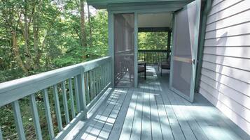 A wooden deck with a railing, leading to a covered porch with a door open, surrounded by trees.