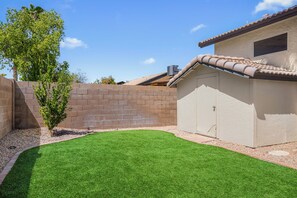 A backyard with a green lawn, a brick wall, and a building with a tiled roof. - Spacious Retreat w/ Free Heated Pool – Close to Stadium & Ballparks! (Peoria)