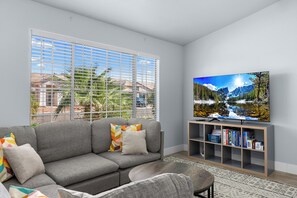 A living room with a grey sofa, colorful pillows, a flat-screen TV, and a bookshelf. - Spacious Retreat w/ Free Heated Pool – Close to Stadium & Ballparks! (Peoria)