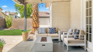 Un patio al aire libre con un sofá color beige, un sillón blanco y una mesa cuadrada con hogar a le ña.
