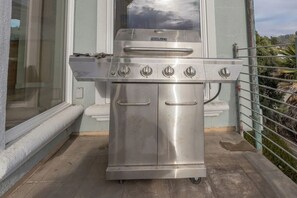 Ein Edelstahl-Barbecue-Grill mit vier Brennern, einem Griff und einem Einstellknopf, platziert auf einem Balkon mit Geländer und Fenster.
