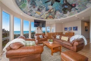 Un salon spacieux avec des canapés en cuir, une table basse et un grand téléviseur. La pièce est dotée d’un plafond haut orné d’une fresque, de grandes fenêtres offrant une vue sur la mer et d’œuvres d’art accrochées aux murs.