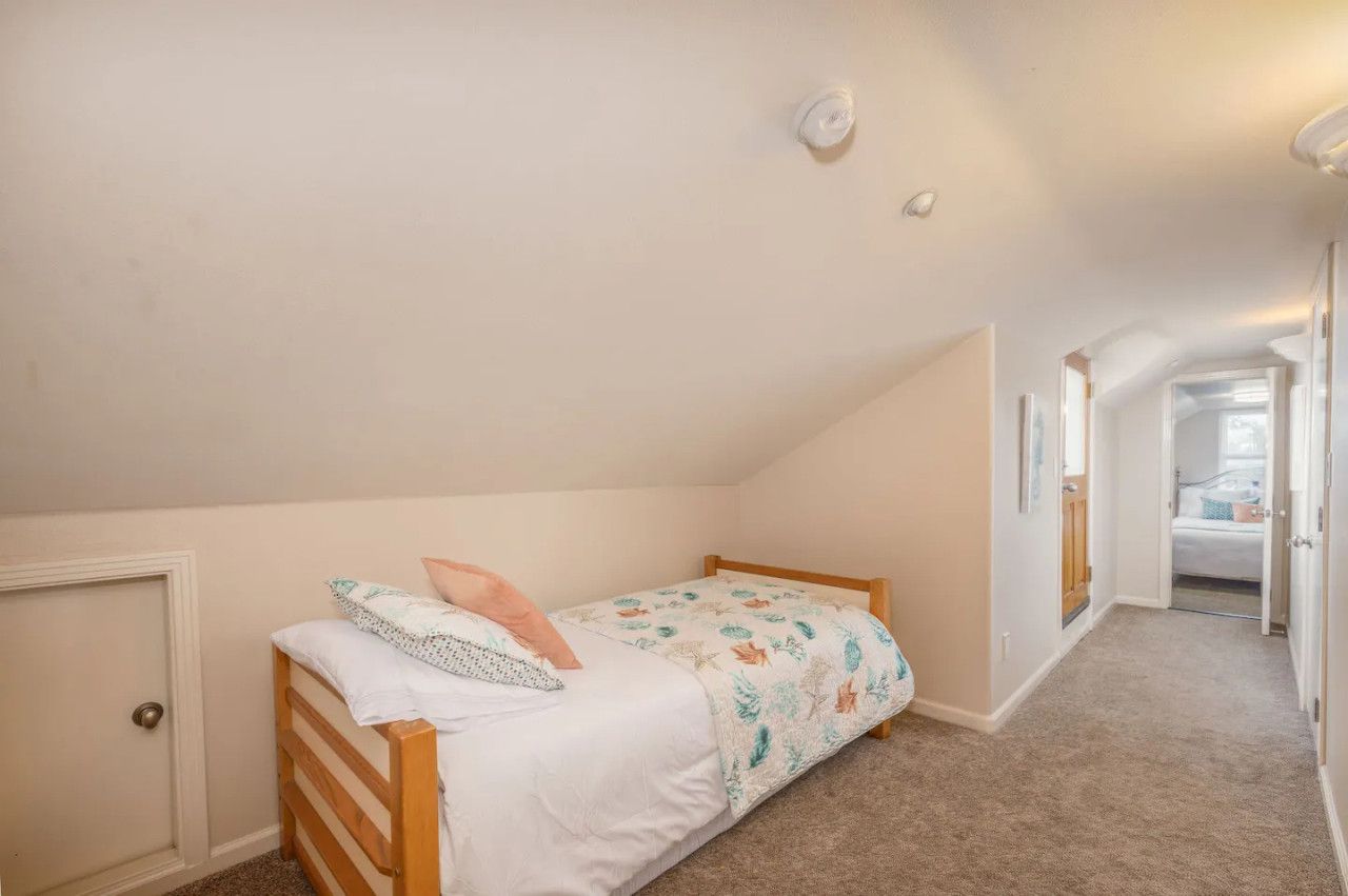 Ein kleines Zimmer mit einem Einzelbett, geneigter Decke, einer Tür links und einem Flur, der zu einem weiteren Zimmer führt.