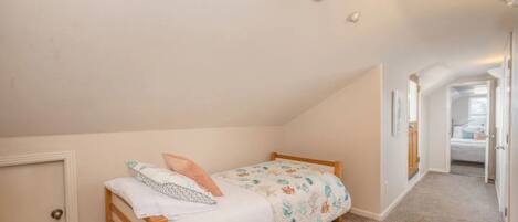 A small, single-bed room with a sloped ceiling, a door on the left, and a hallway leading to another room.