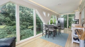 A modern kitchen with a large island, a dining area with a table and chairs, and a view of a lush garden through large windows. - Seaside Escape: 1 block to Beach, Private Hot Tub, Fire Pit & BBQ (El Granada)