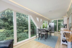 A modern kitchen with a large island, a dining area with a table and chairs, and a view of a lush garden through large windows. - Seaside Escape: 1 block to Beach, Private Hot Tub, Fire Pit & BBQ (El Granada)