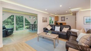 A spacious living area with a large window, a dark leather sofa, a wooden coffee table, and a kitchen in the background. - Seaside Escape: 1 block to Beach, Private Hot Tub, Fire Pit & BBQ (El Granada)