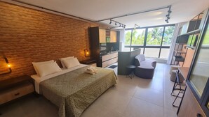 A modern bedroom with a brick wall, a bed with a green bedspread, a small desk, and a large window offering a view of greenery. - URBAN TAMBAU Flat by Penareia.tur.br (João Pessoa)