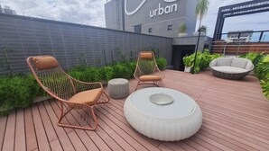 A rooftop patio with wicker chairs and a round table, surrounded by greenery and a modern building in the background. - URBAN TAMBAU Flat by Penareia.tur.br (João Pessoa)