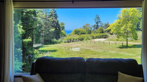 A room with a large window offering a view of a grassy area and trees. - Crescent Beach Retreat: Cozy 4br House (Port Angeles)