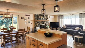 A cozy living room with a fireplace, a dining area with wooden furniture, and a large window offering a view of the outdoors. - Crescent Beach Retreat: Cozy 4br House (Port Angeles)