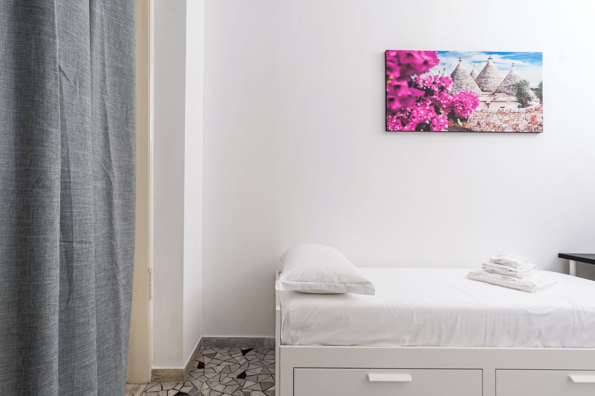 Ein ordentlich bezogenes Bett mit weißen Leinen und Kissen, ein kleiner Nachttisch mit einem gefalteten Handtuch und ein gerahmtes Bild eines Gebäudes mit rosa Blumen an der Wand.