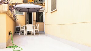 Eine kleine Terrasse im Freien mit Tisch und Stühlen, ein grüner Gartenschlauch und ein weißer Sonnenschirm.