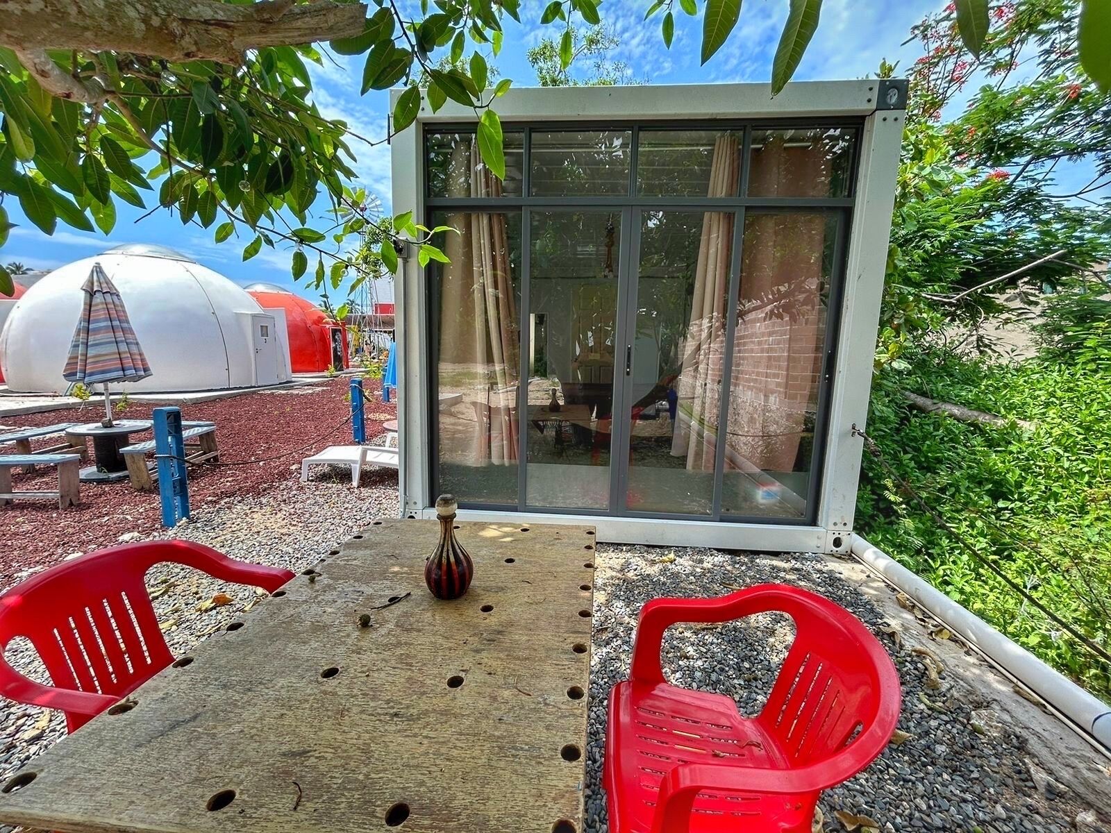 Un petit espace extérieur aménagé avec une table et deux chaises en plastique rouge, un vase décoratif et une porte vitrée donnant sur une pièce.