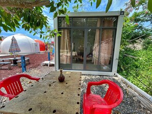 Un'area relax esterna con un tavolo e due sedie di plastica rossa, un vaso decorativo e una porta in vetro che dà su un ambiente interno.