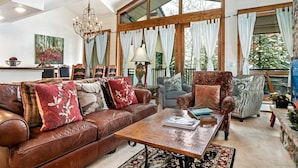 A spacious living room with a leather sofa, patterned armchairs, and a coffee table. Large windows with curtains allow natural light and views of greenery outside. - Antlers Condominiums - Vail (Vail)