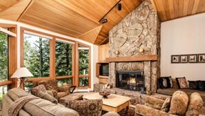 A cozy living room with a stone fireplace, wooden ceiling, and large windows offering a view of trees. - Snowmass Woodrun V Residences (Snowmass Village)