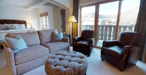 Une chambre d’hôtel comprenant un lit, un canapé, deux fauteuils en cuir, un pouf à motifs et un balcon avec vue sur les montagnes.