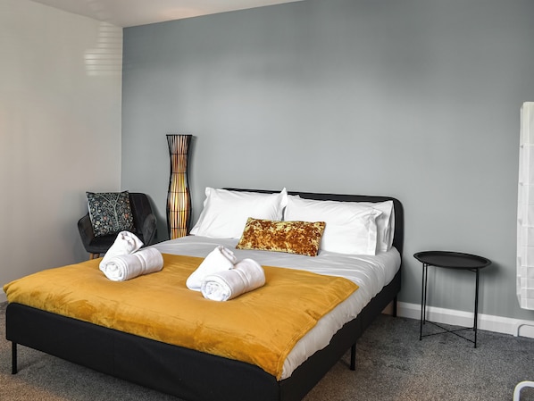 A neatly made bed with white linens and a mustard-colored throw, a tall floor lamp, a black chair, and a small side table.