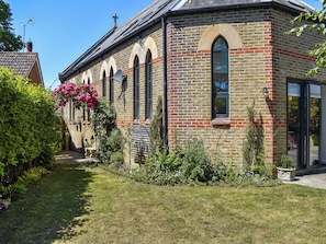 A brick building with arched windows and a cross on the roof, surrounded by a well-maintained garden.