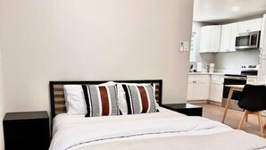 A modern bedroom with a bed, a nightstand, a chair, and a kitchenette in the background. - Stylish Heaven/Central SA (San Antonio)
