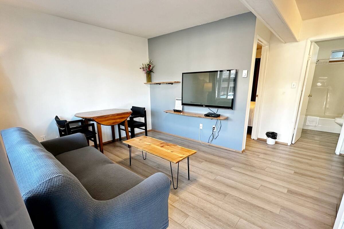 A modern living room with a sofa, a wooden coffee table, a TV mounted on the wall, and a small dining table with chairs.