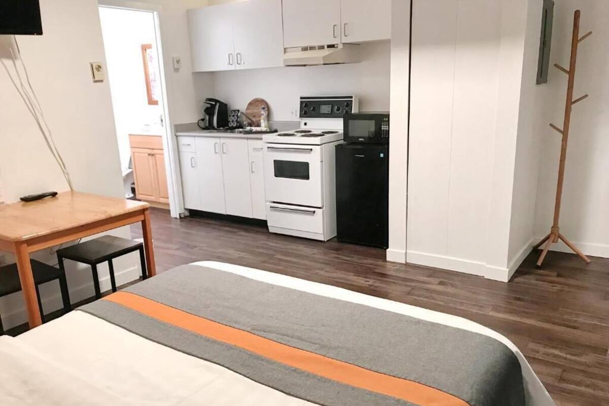 A bedroom with a bed, a wooden table, and a kitchenette with a stove, microwave, and cabinets.