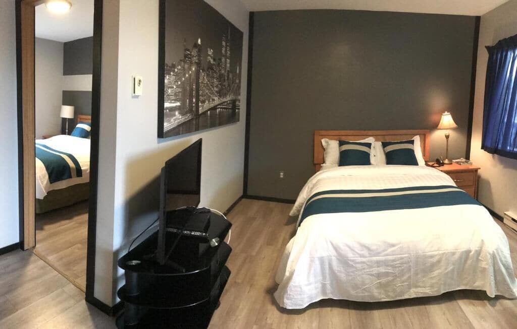 A hotel room with a bed, a TV, a cityscape painting, and a suitcase.