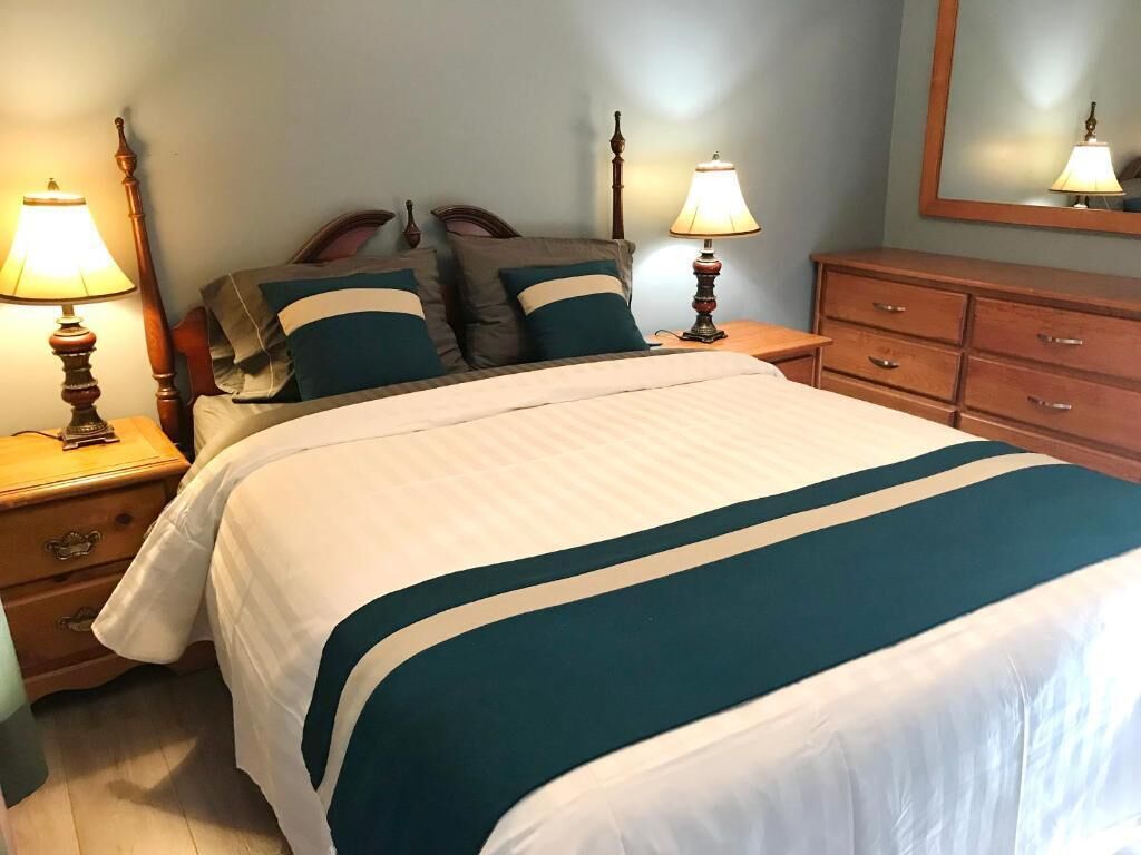 A neatly made bed with a white and teal bedspread, flanked by two bedside tables with lamps, a wooden dresser, and a mirror on the wall.