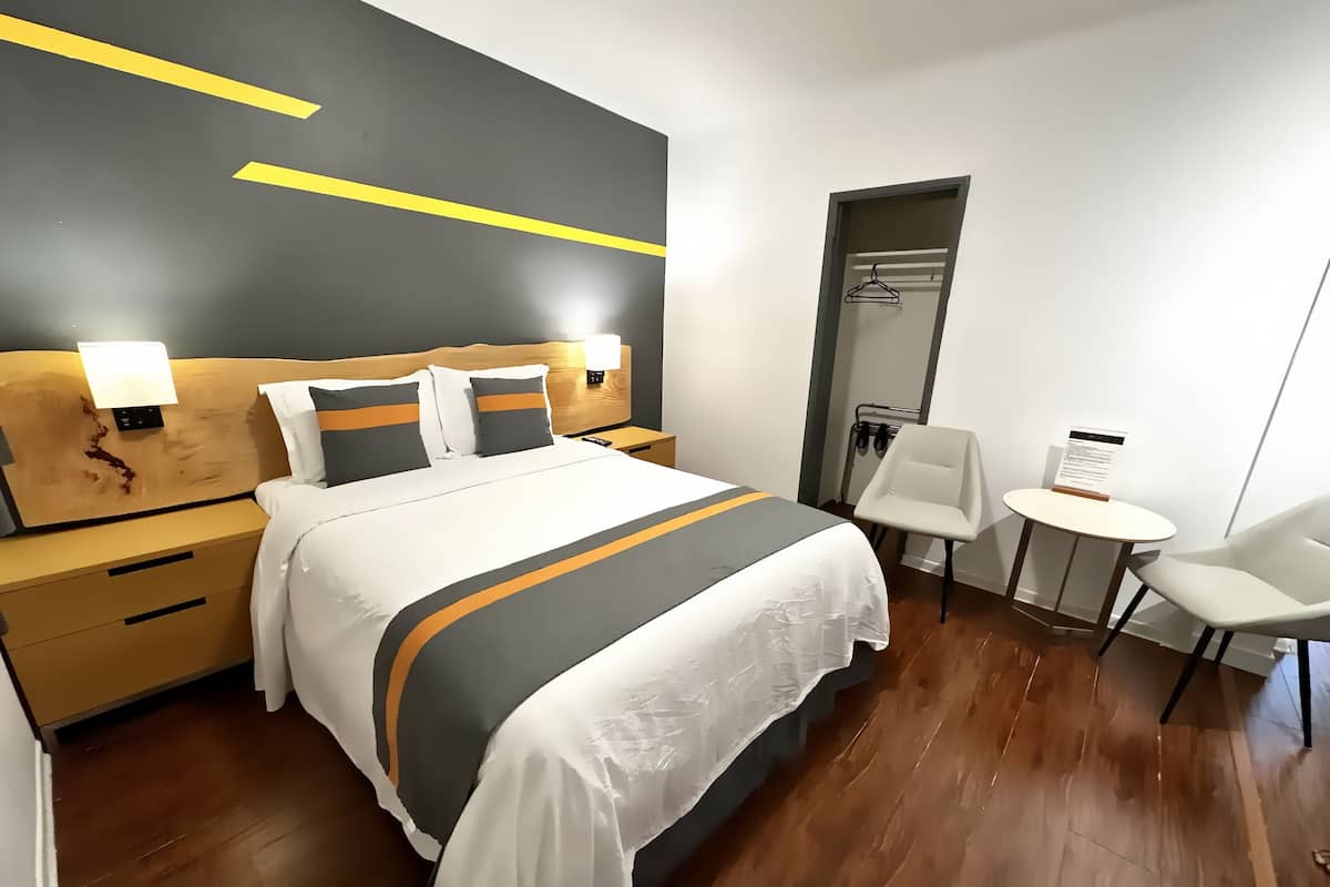 A modern hotel room with a large bed, bedside tables, a small round table, and a chair.