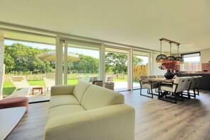 Amplia sala de estar con un sofá grande, mesa de comedor y sillas. Grandes ventanales ofrecen vistas al jardín.