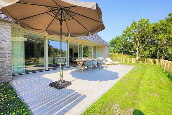 Una casa moderna con un patio, mobiliario exterior y un amplio paraguas.