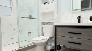 Un baño moderno con ducha envuelta en vidrio, un inodoro, un lavabo con espejo y un mueble con artículos de aseo.