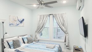 Un dormitorio con cama, ventilador de techo, ventana con cortinas, televisor y escritorio con silla.