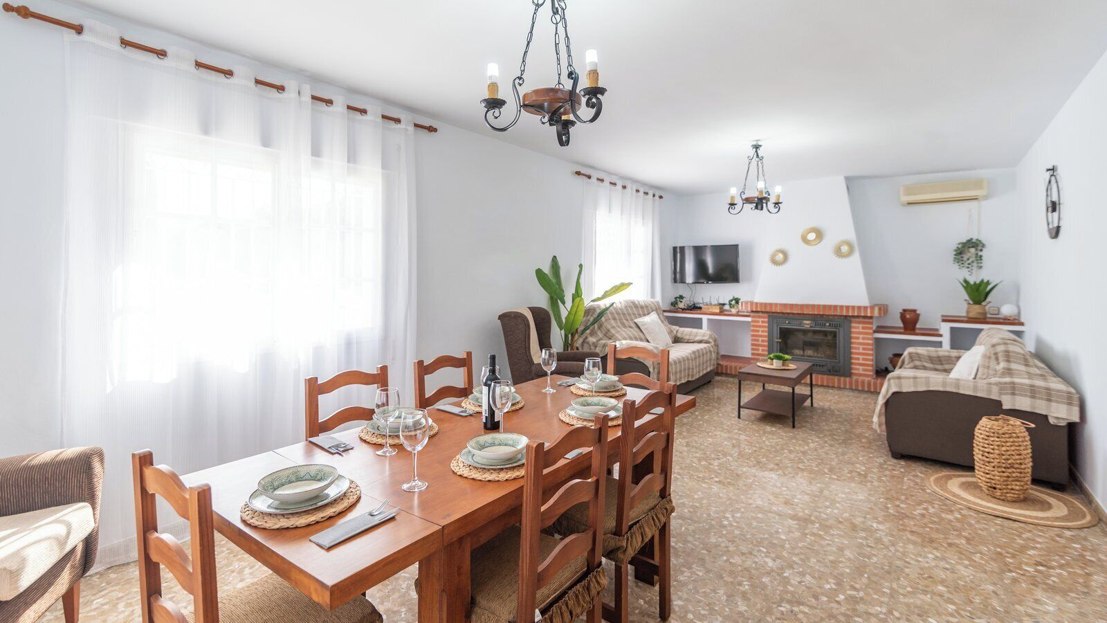 Uma área de jantar com mesa de madeira posta para quatro pessoas, cercada por cadeiras. Há um sofá e uma pequena mesa com uma planta ao fundo.