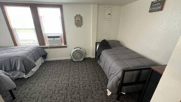A small, compact bedroom with two single beds, a standing fan, a window with blinds, and a small wall-mounted heater.
