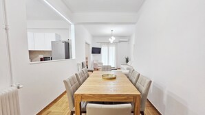 Un espace repas moderne avec une table en bois et des chaises rembourrées, une cuisine avec des meubles blancs et un téléviseur fixé au mur.
