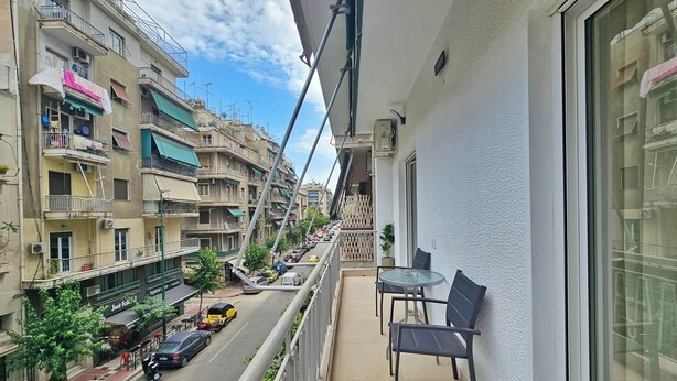 Un balcon avec une table et des chaises, donnant sur une rue bordée de voitures garées et d’immeubles.