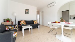 A modern living room with a black sofa, a white coffee table, and a dining area with a white table and chairs. There is a ceiling fan, an air conditioning unit, and a kitchen area with a refrigerator and a microwave. - CT 288 AL   Faro's Retamar Cuddly Nest for 2 (Almería)
