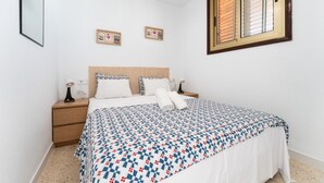 A bedroom with a bed, two bedside tables, a nightstand, a window with blinds, and two wall-mounted decorations. - CT 288 AL   Faro's Retamar Cuddly Nest for 2 (Almería)