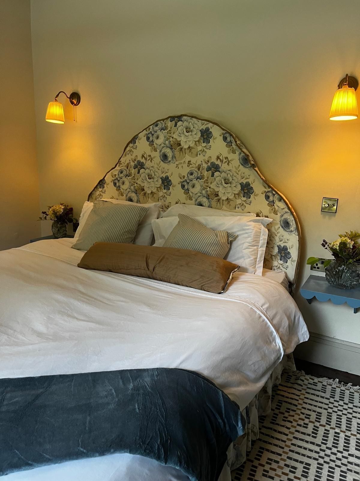A neatly made bed with a floral headboard, two bedside lamps, and a small table with a vase of flowers.