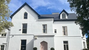 A two-story white building with a symmetrical facade, a central entrance with a pink door, and a small garden area with potted plants and outdoor furniture. - Lynwood house (Cobh)