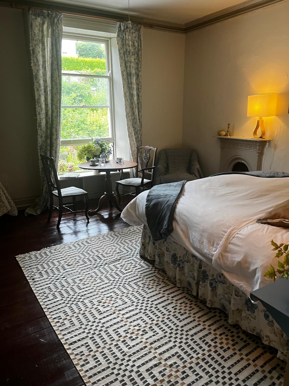 Un dormitorio con una cama, una mesita con sillas, una ventana con cortinas y una chimenea con una lámpara.