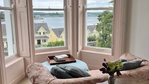 A bay window with a view of a river and houses, a sofa with a floral pattern, and a small table with a vase of flowers. - Lynwood house (Cobh)