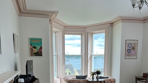 A hotel room with a piano, a floral sofa, a bed, and a view of the water. - Lynwood house (Cobh)