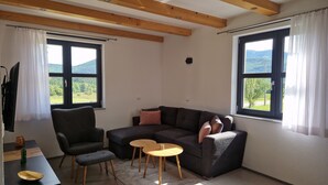 Una sala moderna con un sofá seccional gris oscuro, una pequeña mesa redonda de madera, un sillón gris y un mueble bajo para la televisión negro. Hay dos ventanas con cortinas blancas, una de las cuales ofrece vistas a zonas verdes y colinas.