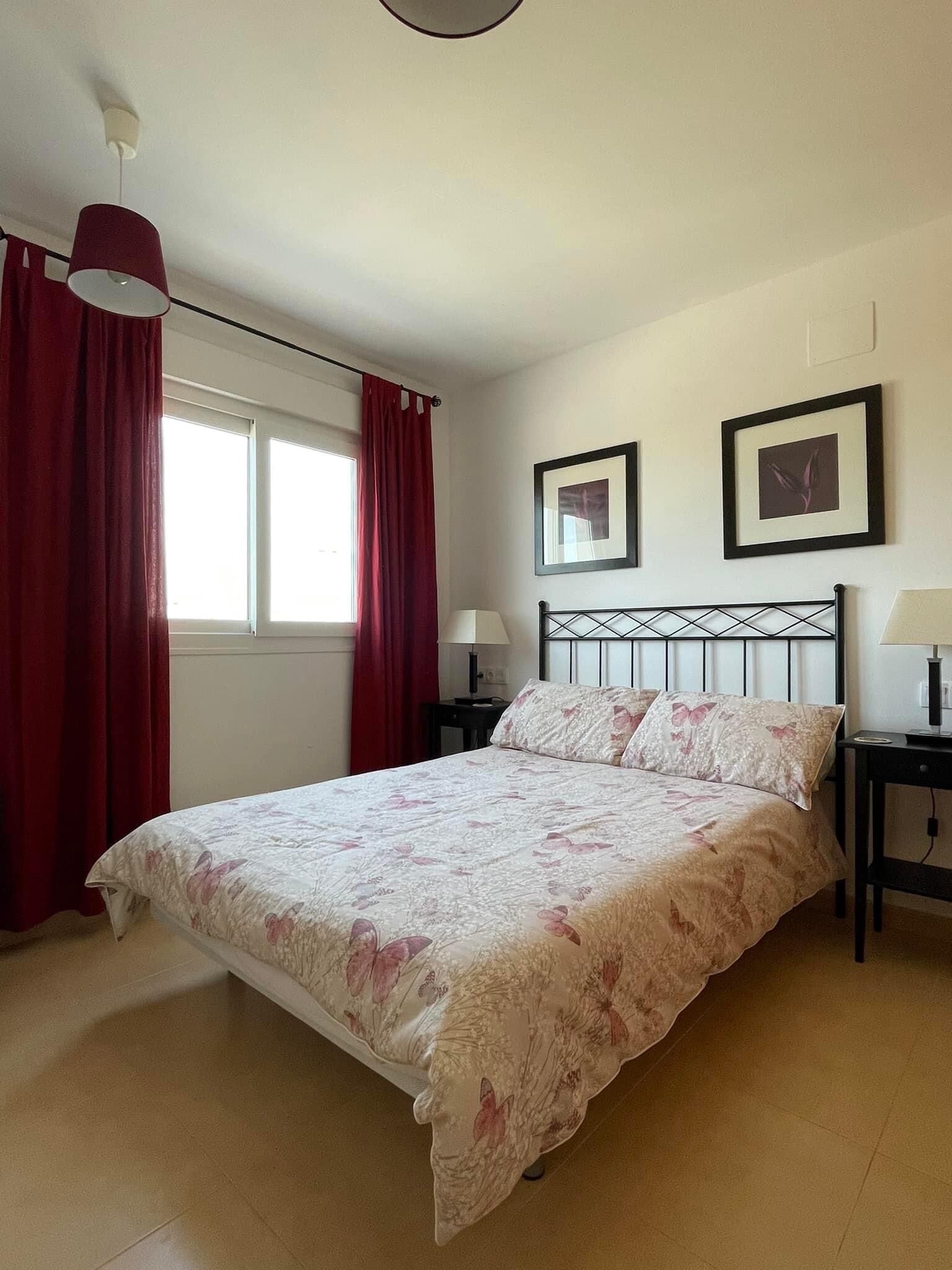 Une chambre à coucher avec un lit, deux tables de chevet, une fenêtre avec des rideaux rouges et deux tableaux encadrés accrochés au mur.